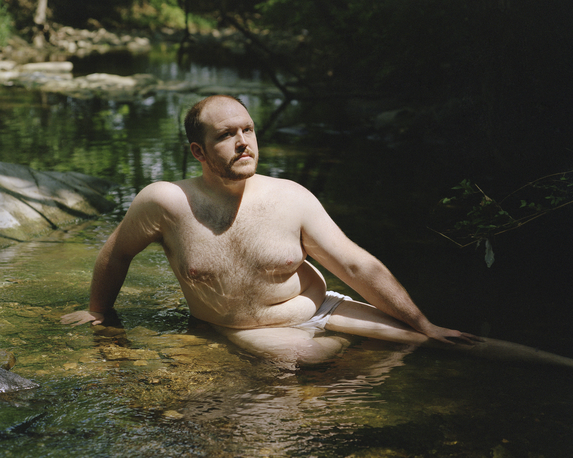 a person sits in their underwear in a rocky stream, looking at the camera
