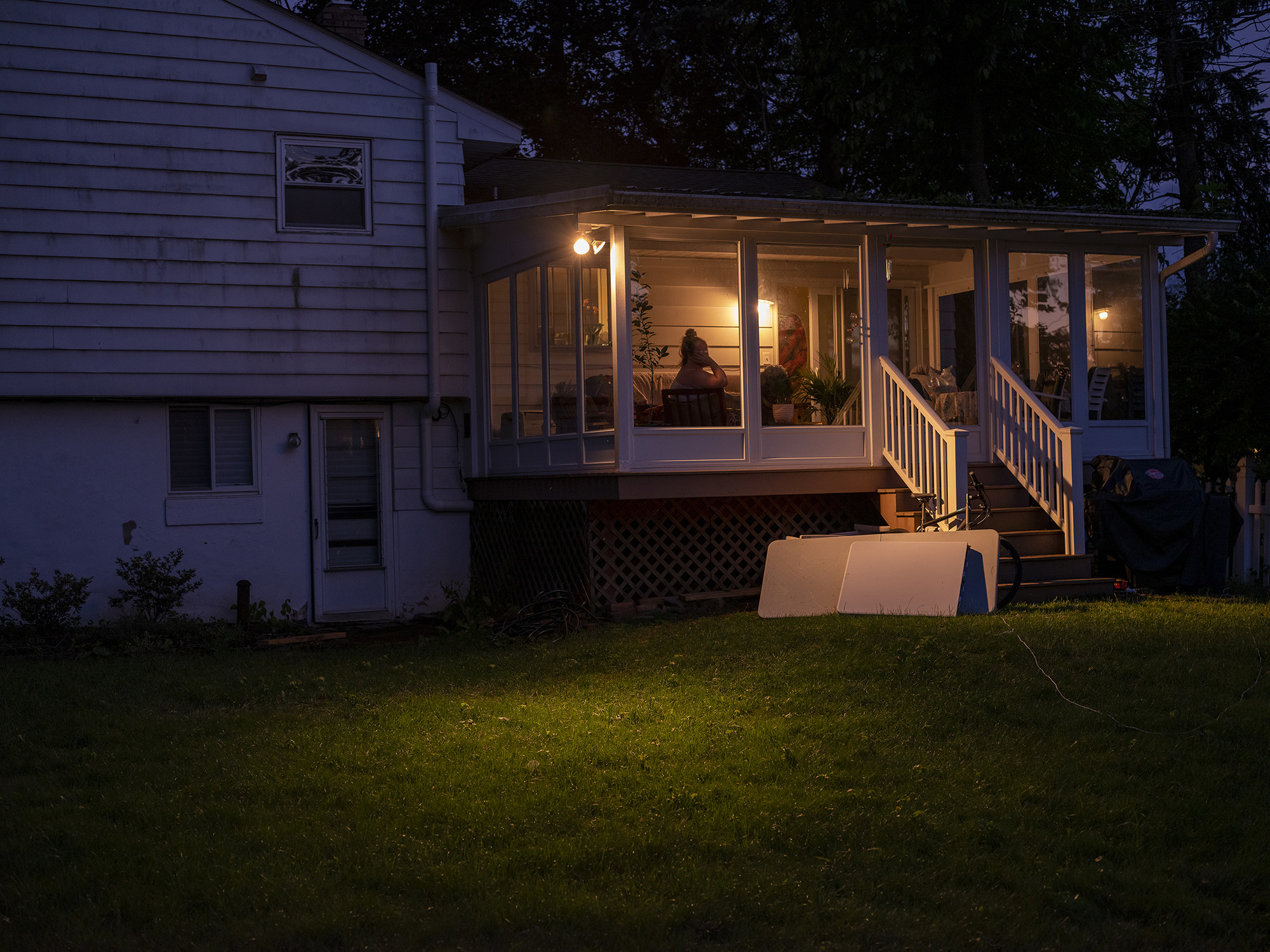 A shot of a person sitting in a glass enclosed porch behind their home, with a porch light on 