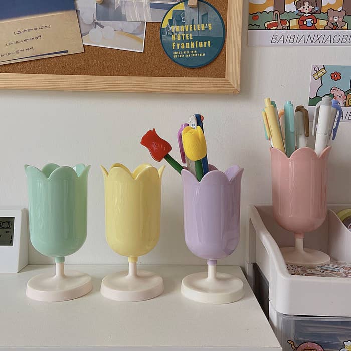 four tulips pencil cups in green, yellow, purple, and pink, sitting on a desk beside each other.