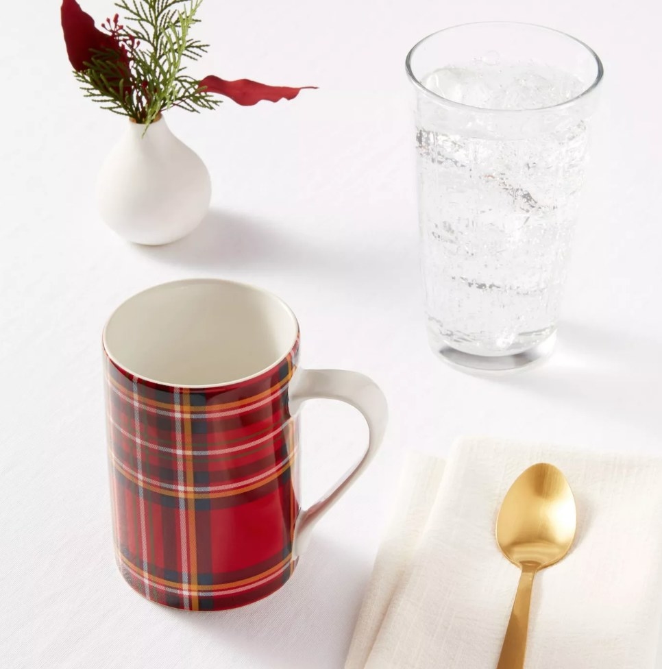 Red plaid mug next to gold spoon on napkin, small vase with red and green plant behind it, tall glass of water on right