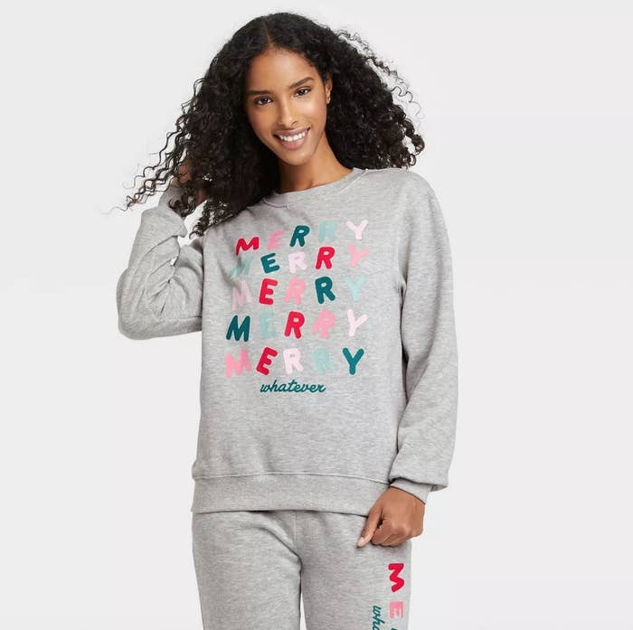 Model wearing gray crewneck that says &quot;Merry Merry Merry whatever&quot; in red, pink and green lettering