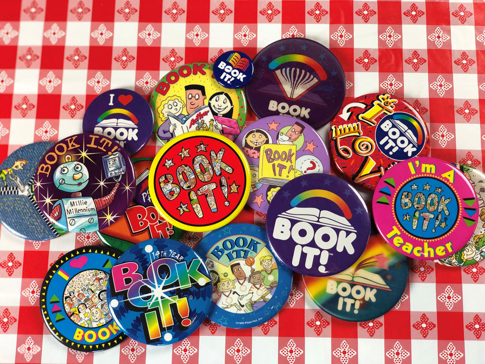 A collection of BOOK IT! buttons with different designs sitting atop a red-and-white checkered tablecloth