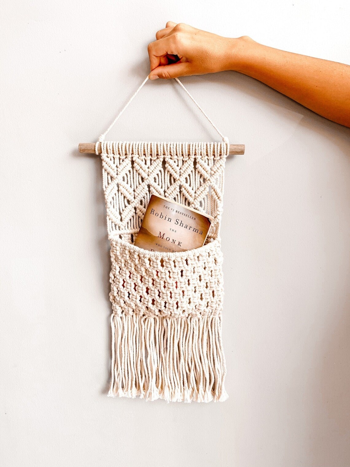 the cream-colored macrame  pouch with a wooden beam holding it at the top for hanging and fringe at the bottom