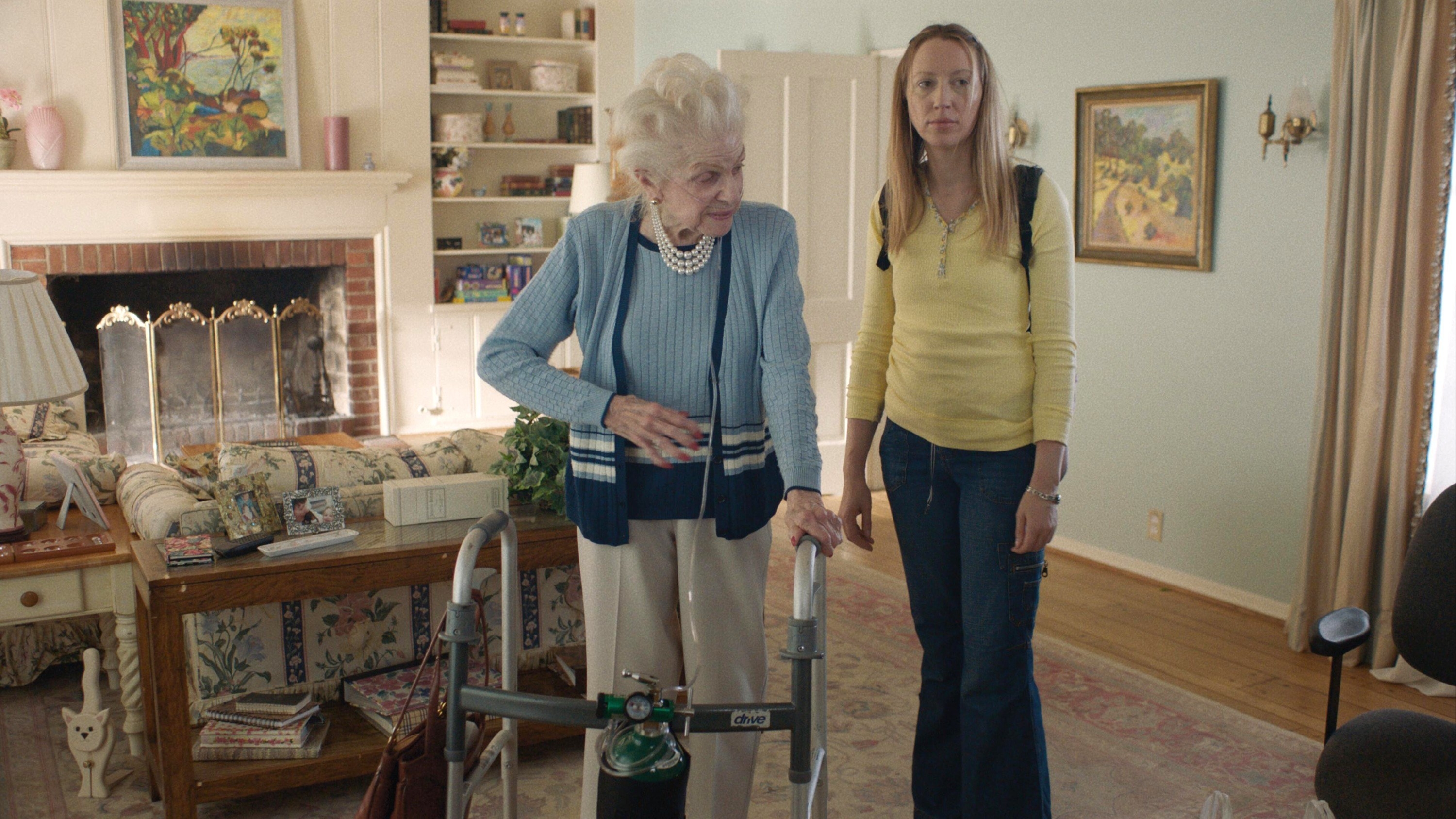 Anna stands, frowning next to her grandma who uses a walker and oxygen tank