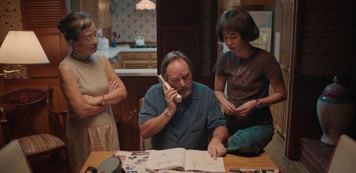 Yuki (mom) stands arms crossed next to dad, seated on the phone, looking at a magazine while Maya looks over his shoulder