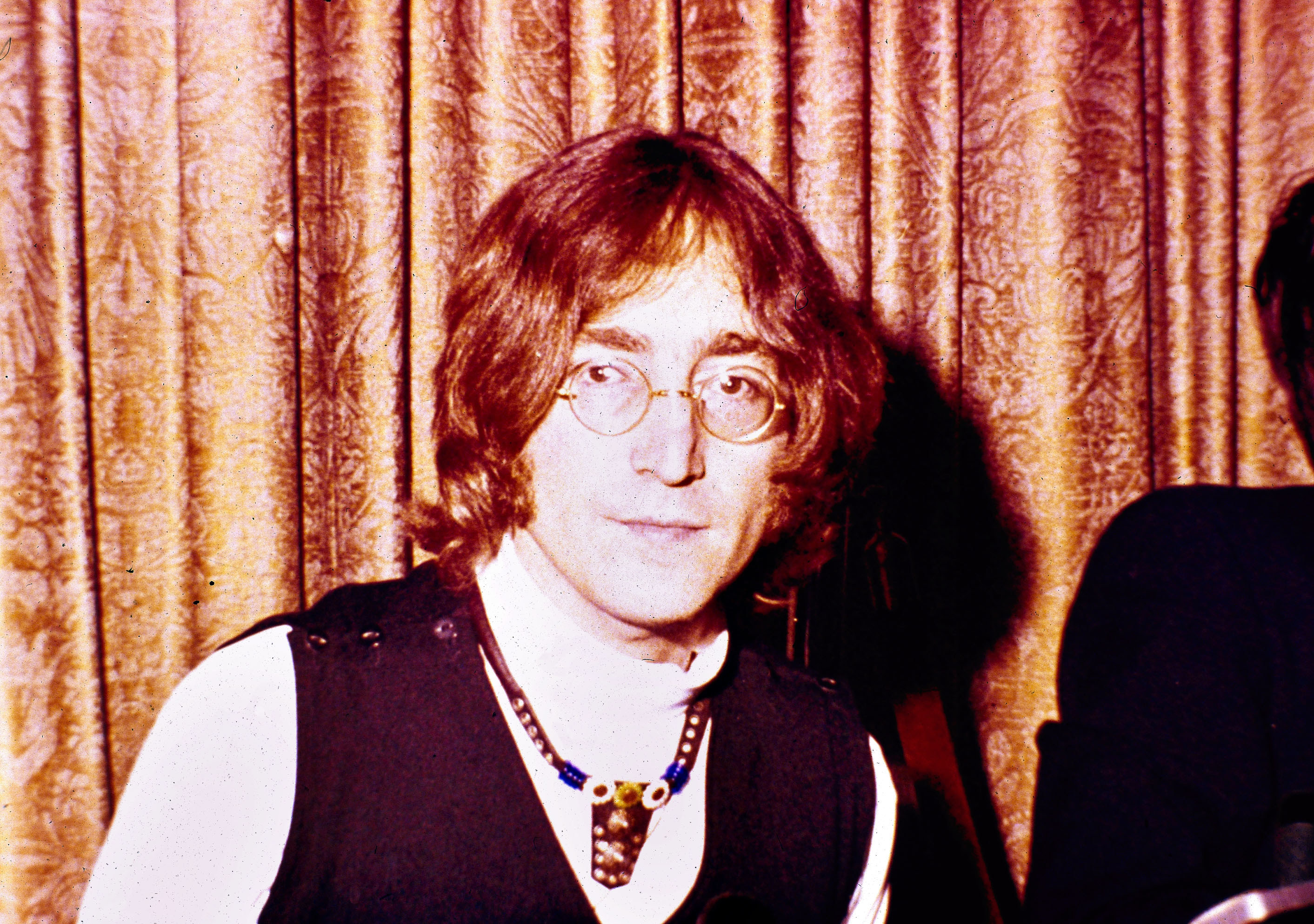 John Lennon in front of a curtain with long hair his signature glasses and a necklace