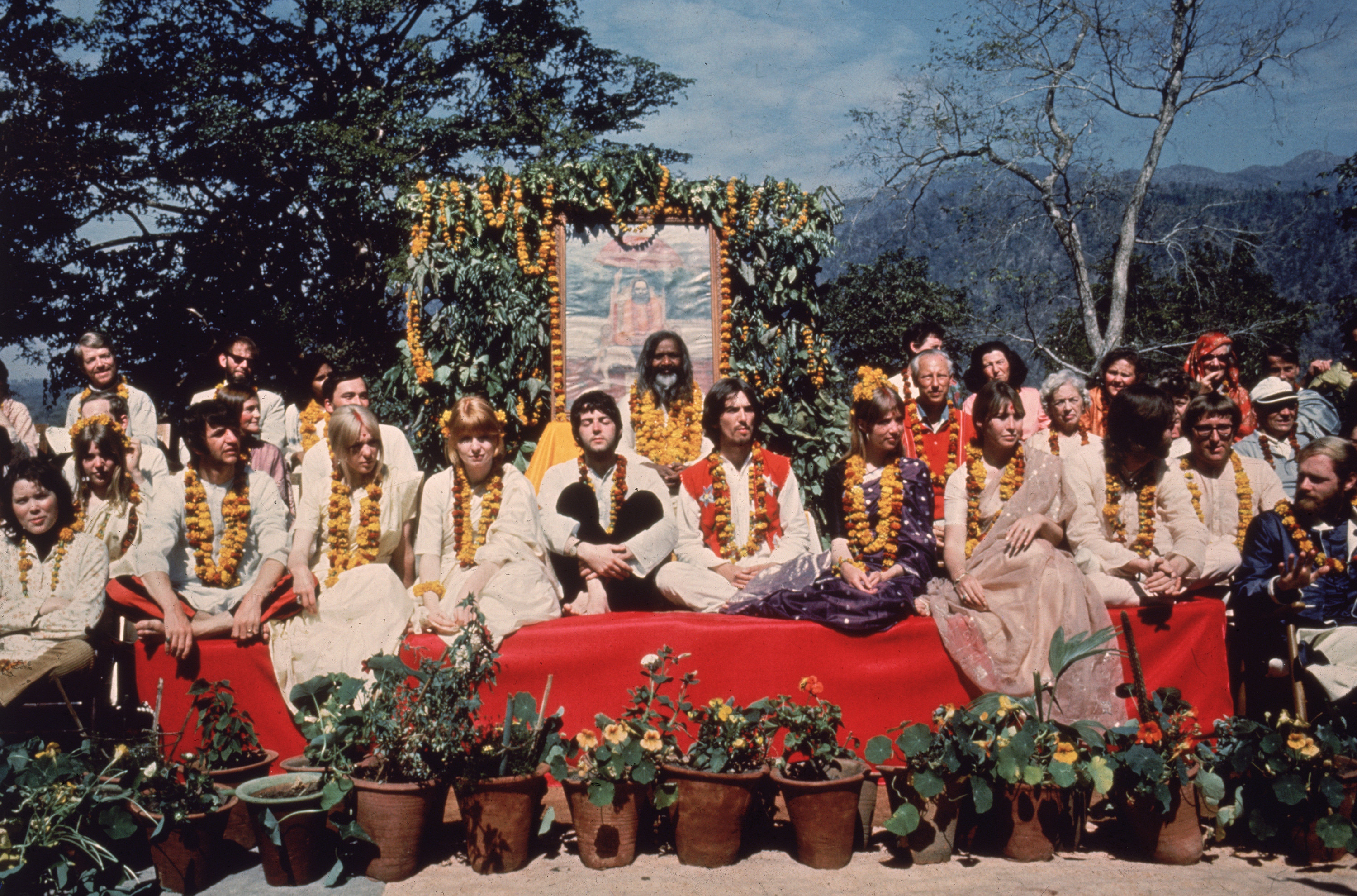 the beatles and their wives sit in a large group of people on a red platform wearing flower wreathes with a guru in india