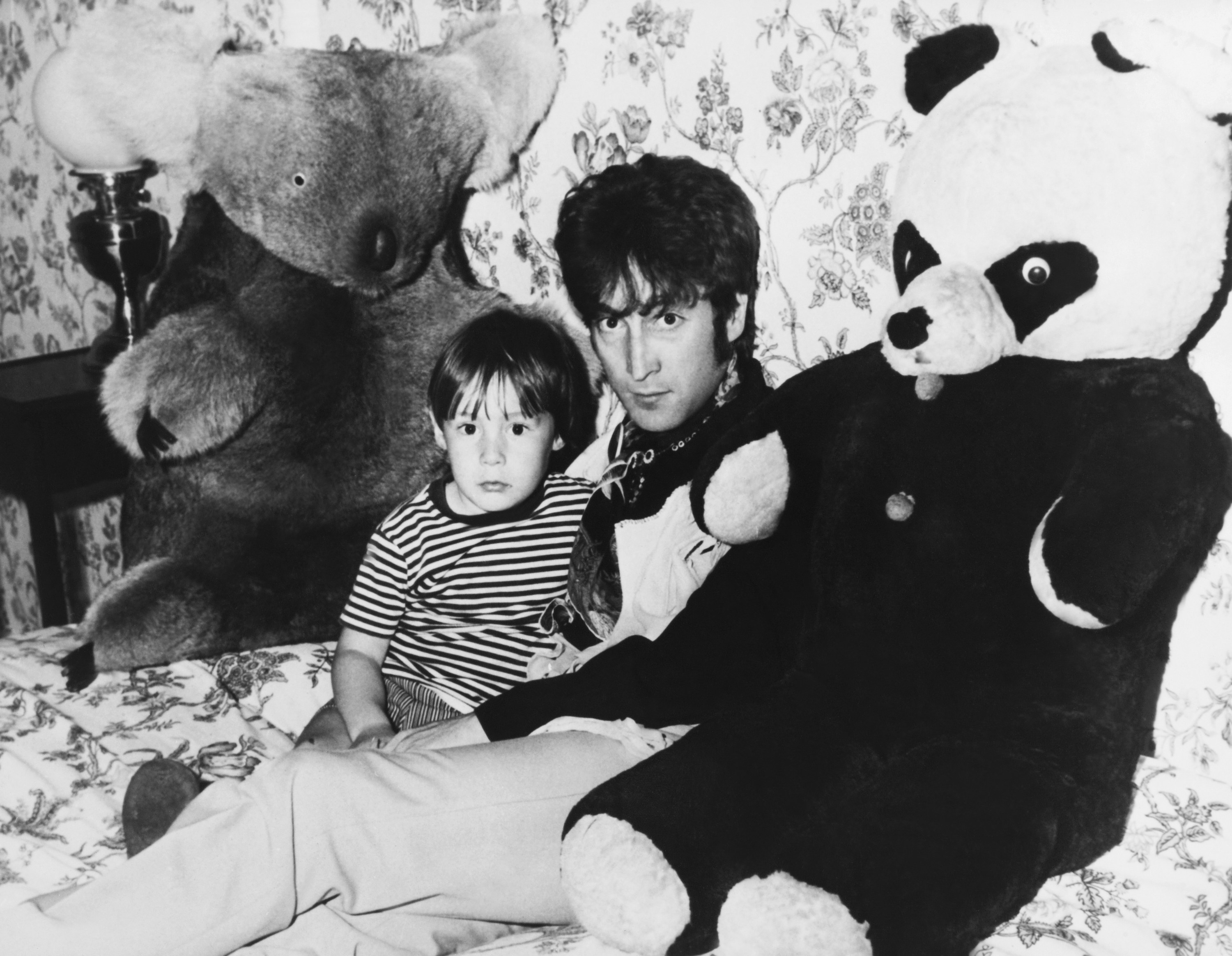 John Lennon and his young son on a bed with floral pattern sheets sitting between two giant stuffed bears