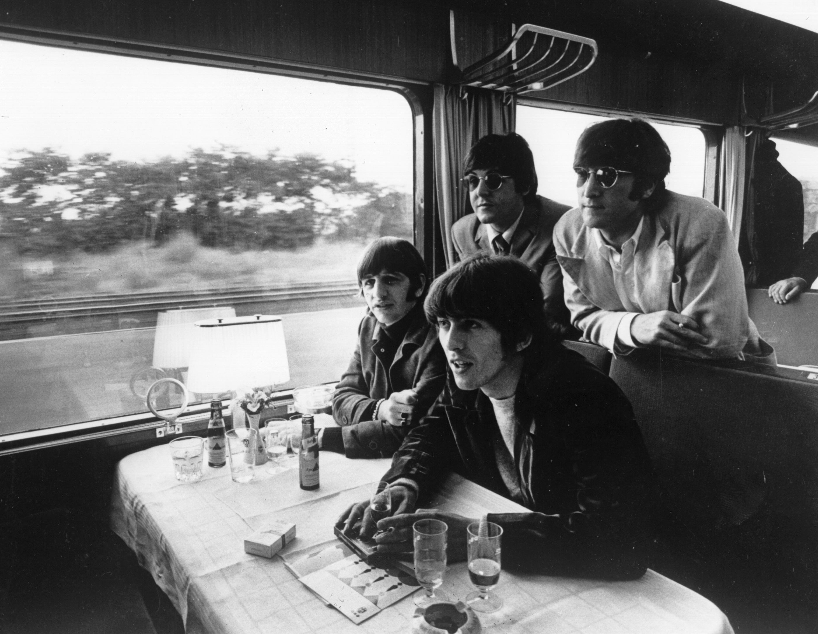 The Beatles on a train in one booth, with glasses beer and cigarettes on the table as the landscape goes by