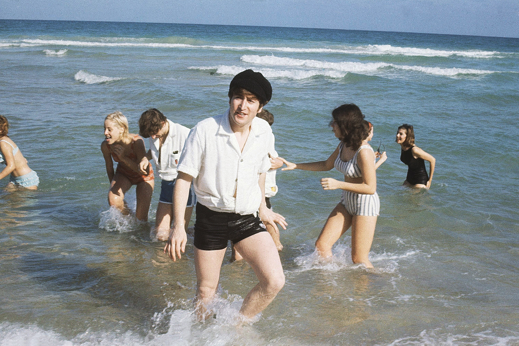 john lennon in shorts and a button down shirt wading in the ocean with a crowd