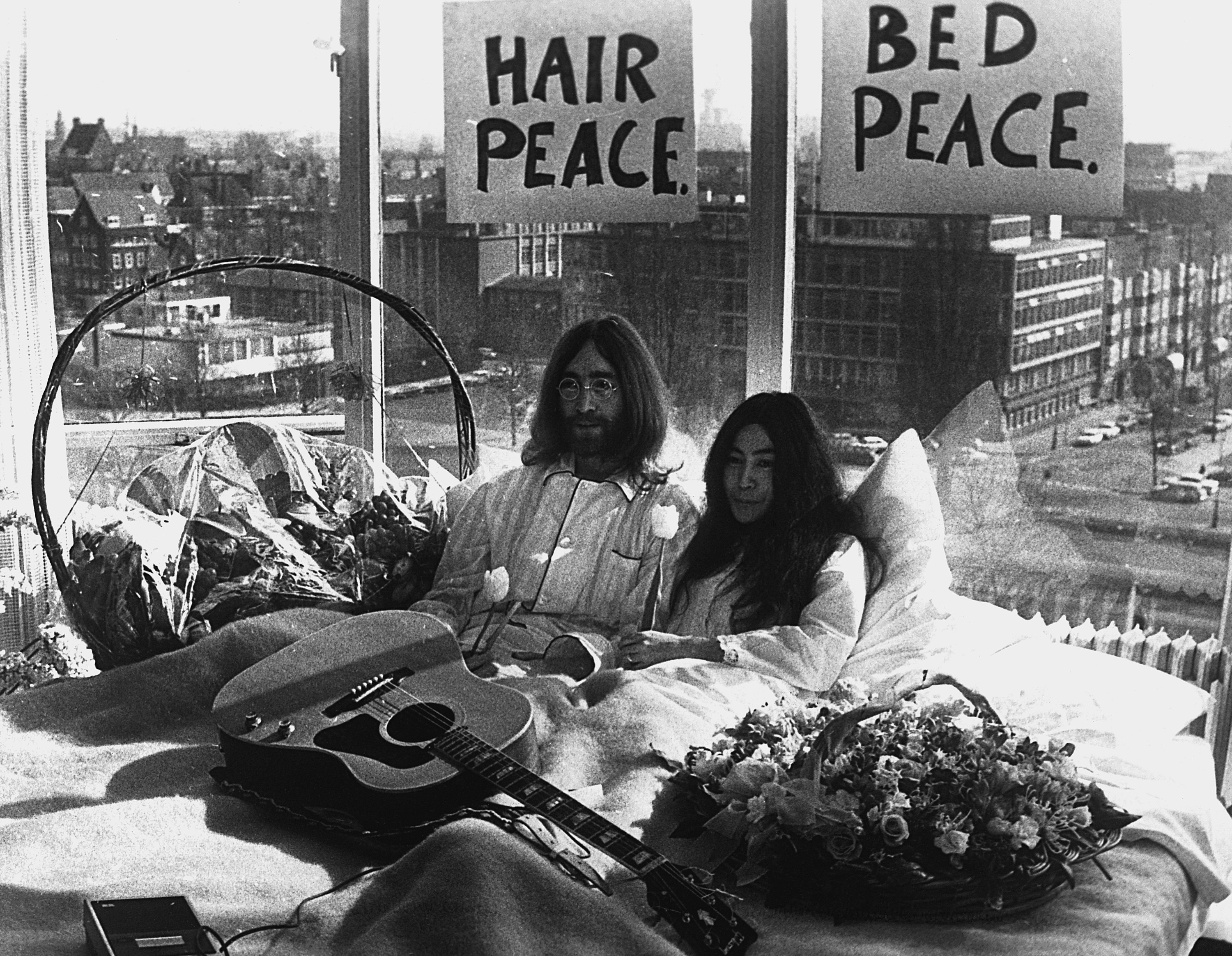 John Lennon and Yoko Ono  in bed with signs behind them that reads hair peace bed peace a guitar and flowers are on the bed