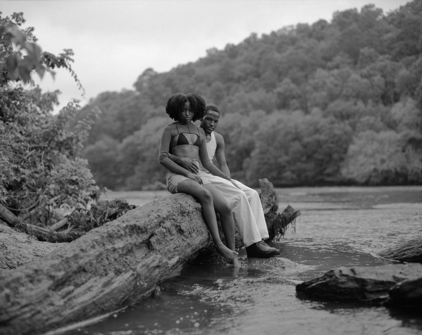 A man and woman sitting on a fallen log by a river with trees in the background