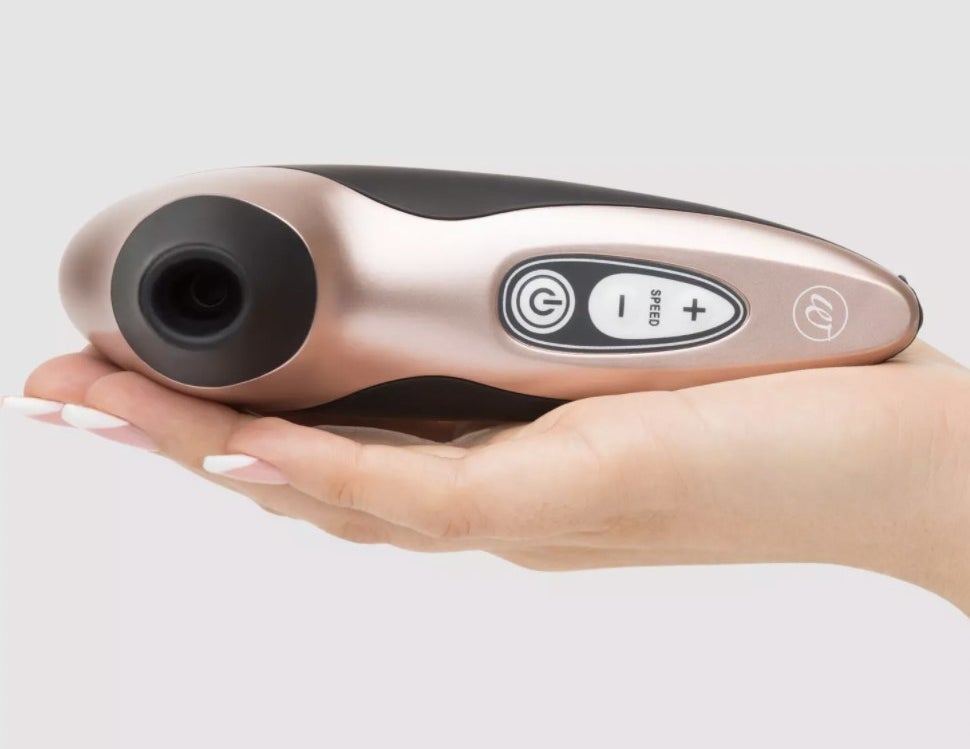 The pink stimulator resting in someone&#x27;s palm, showing the silicon-tipped suction area and control panel, with power button and - and + controls for speed