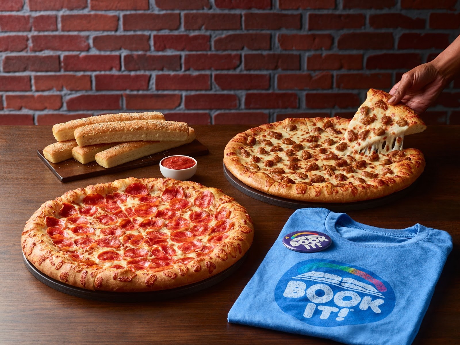 A product shot showing two Pizza Hut pizzas, breadsticks, a BOOK IT! button, and a folded BOOK IT! T-shirt