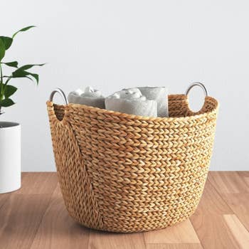 Woven basket with metal handles holding rolled towels, placed on a wooden floor. A small potted plant is visible in the background