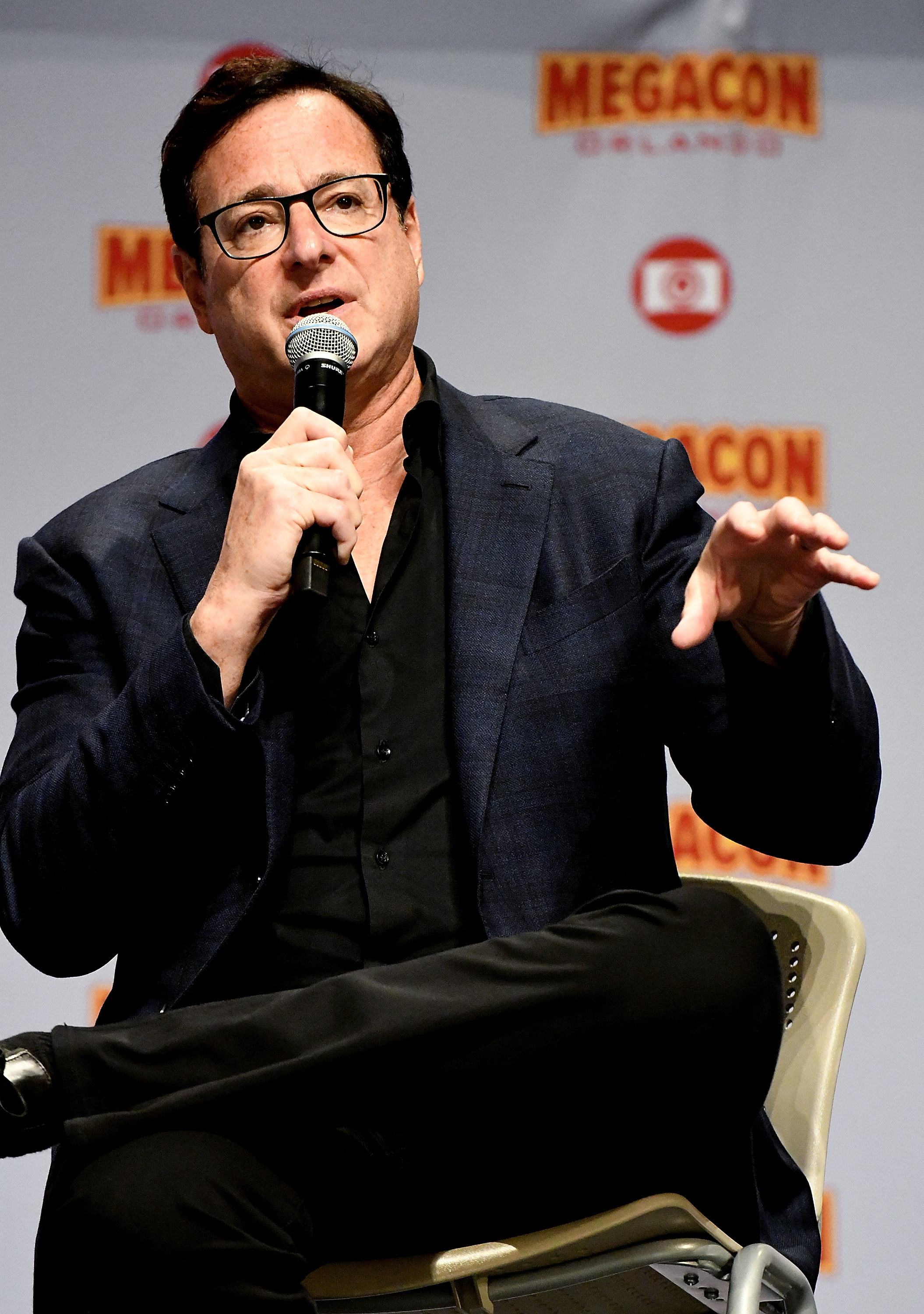 Saget extends his arm while speaking into a microphone