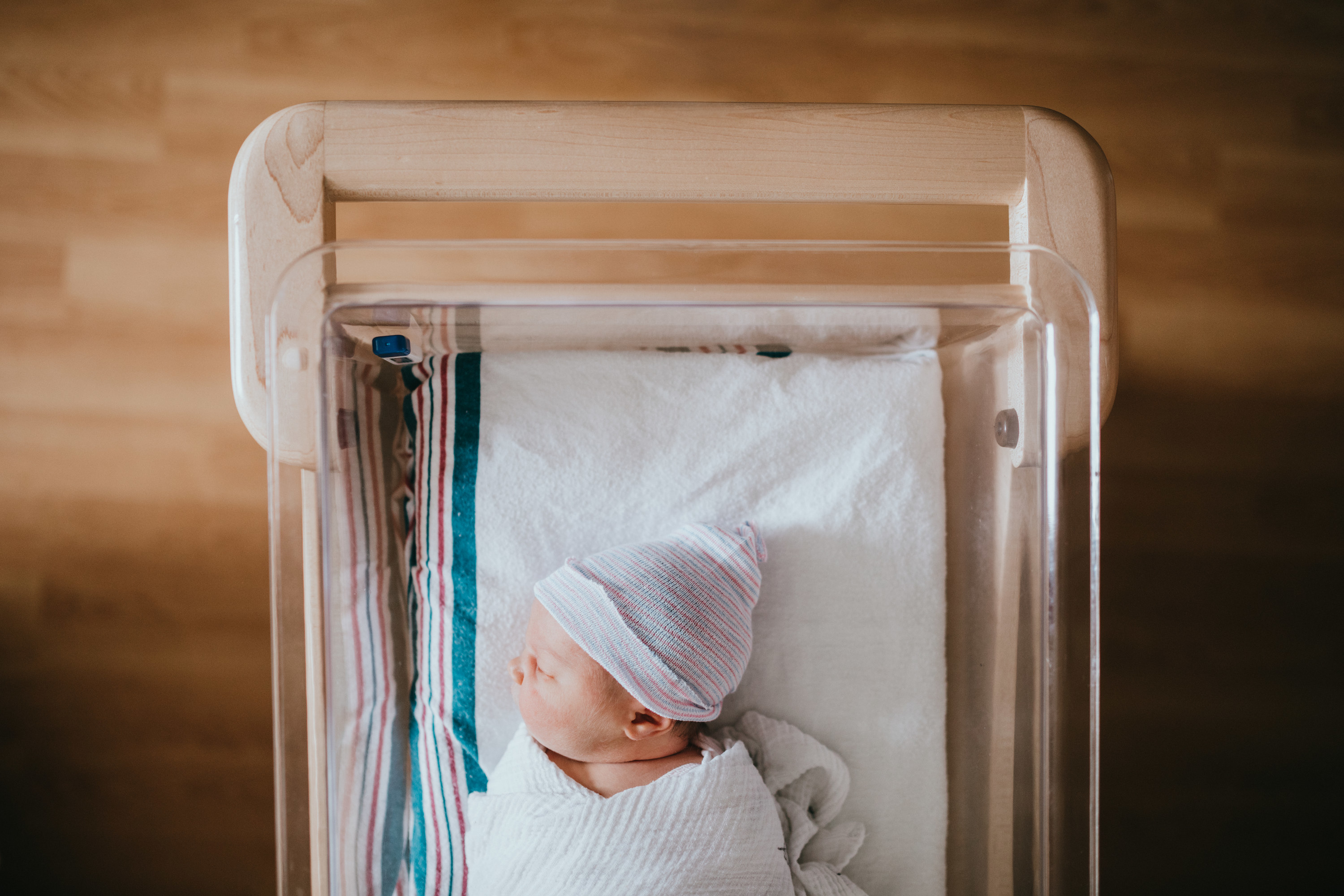 A baby just born at the hospital rests in a hospital bassinet crib, wrapped in a swaddle and wearing a beanie hat