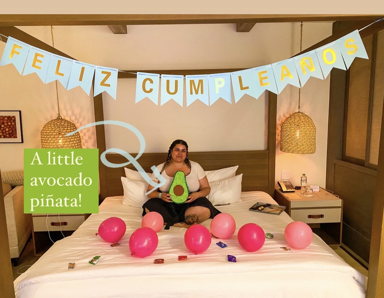 Woman sitting on a bed and holding a little avocado piñata in a room decorated for a birthday, with the sign &quot;Feliz cumpleaños&quot; hanging above the bed and balloons and sweet treats on the bed