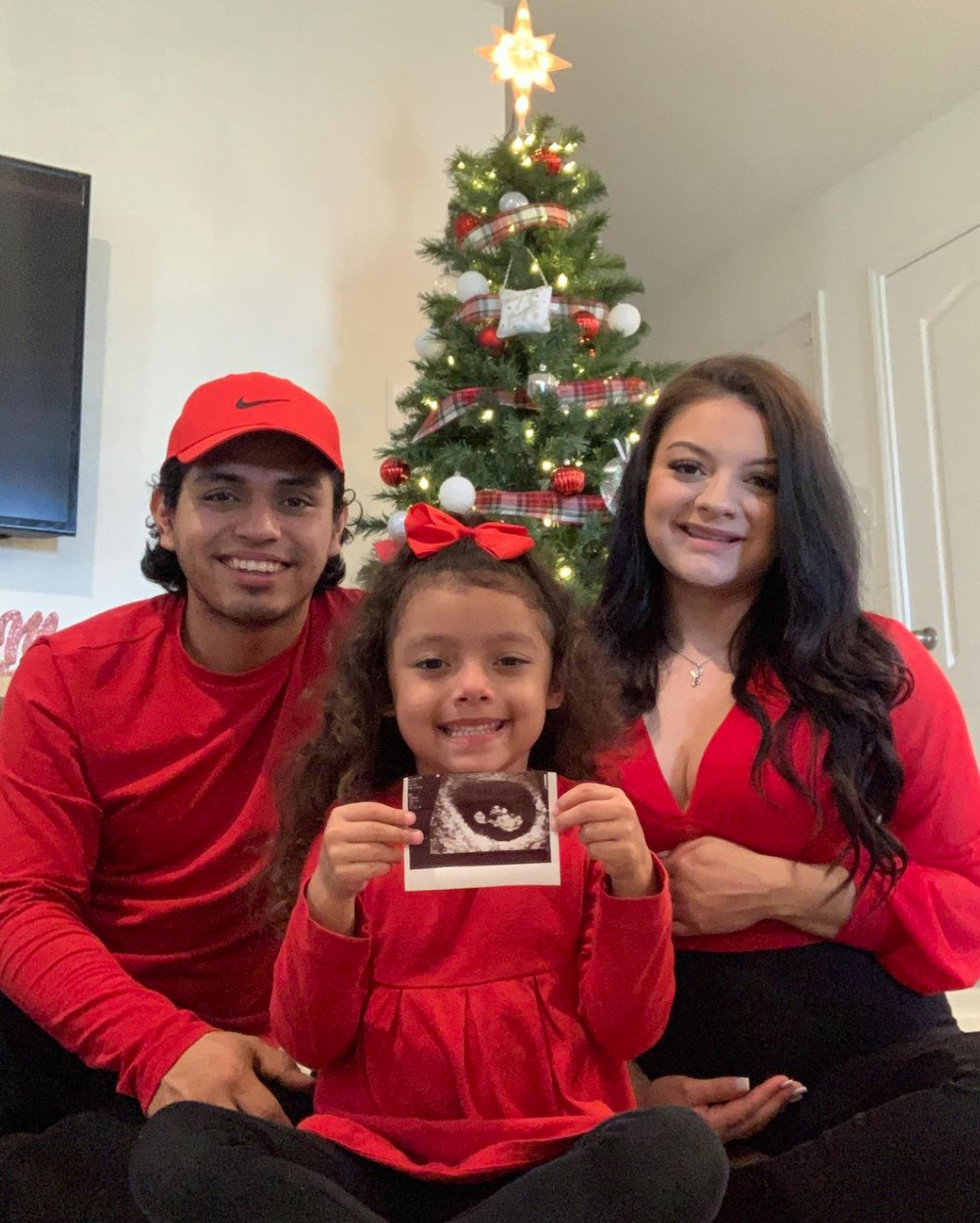 Jasmine and her partner posing for a photo with their daughter who&#x27;s holding up the ultrasound of the baby