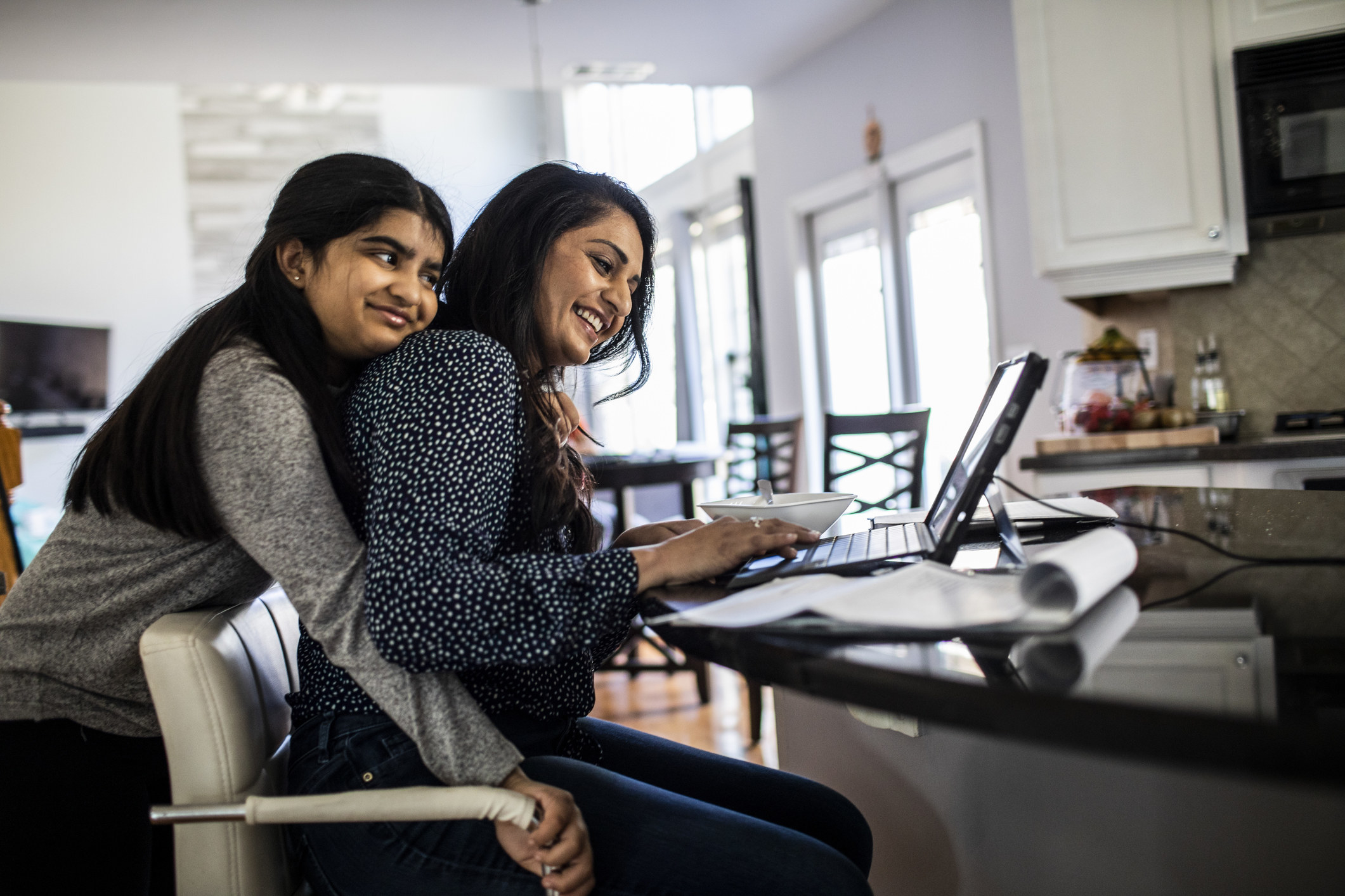 Woman working from home in kitchen while teenager daughter hugs her