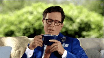 Stephen Colbert sits on a couch and blows steam off of a mug of liquid before inhaling it in