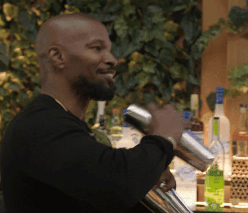 Jamie Foxx smiles and shakes two cocktail mixers in his hand as he sways back and forth in
