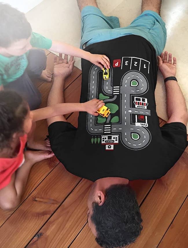 A parent laying on their front on the floor wearing the shirt that has a car race track printed on it while kids play with cars on it