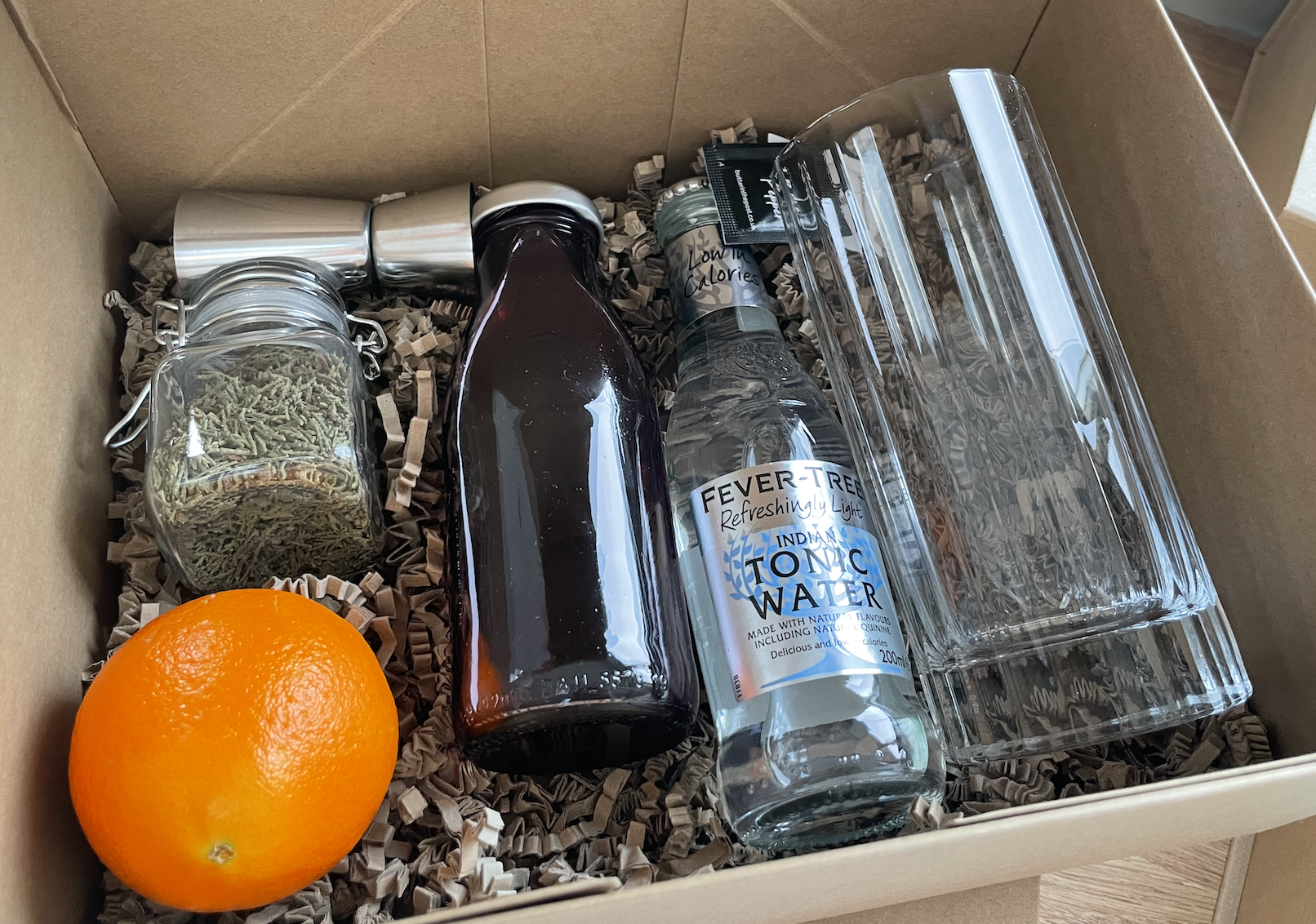 a box filled with ingredients for a winter gin and tonic including an orange juniper sprigs a deep red cordial and a bottle of tonic water