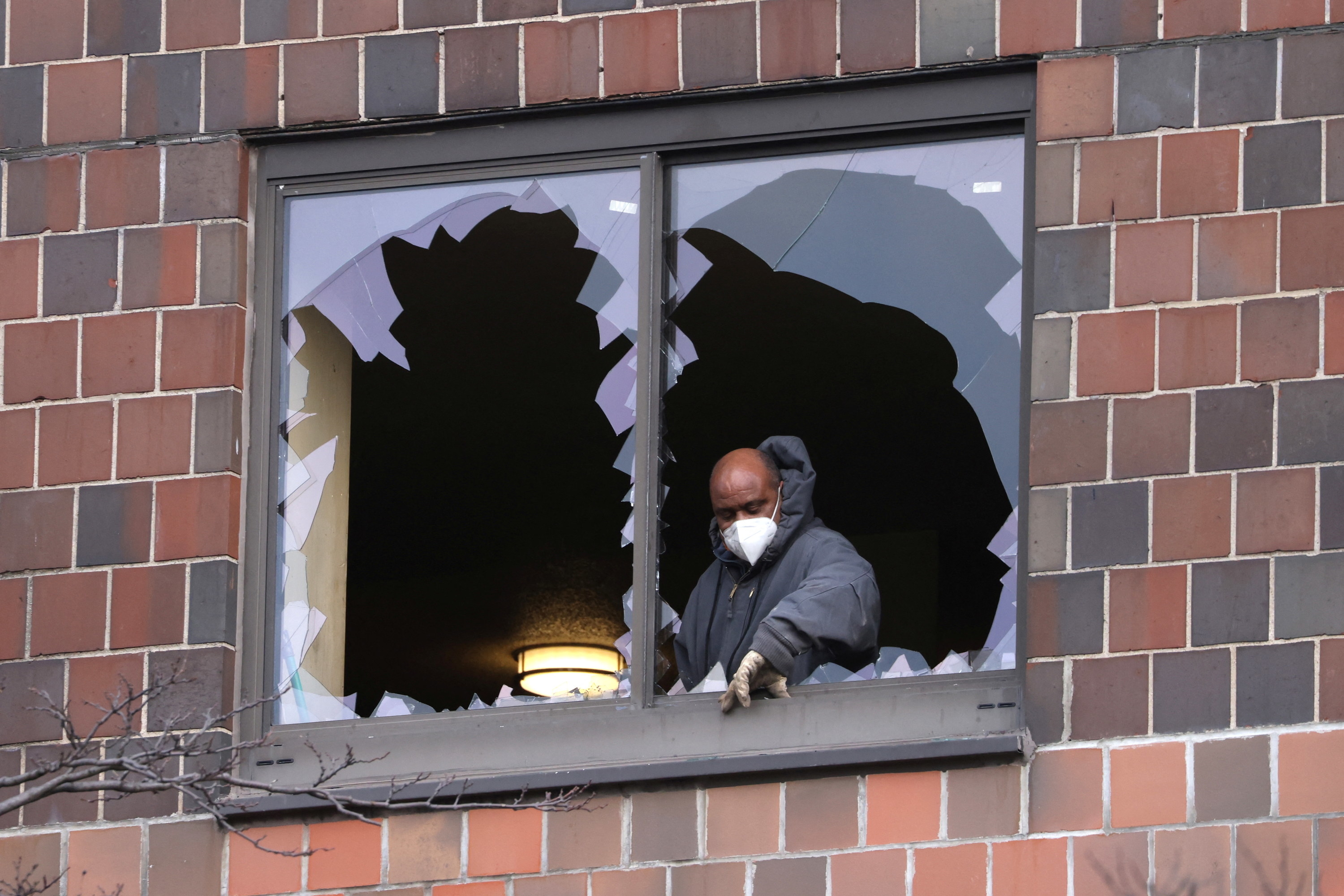 A man wearing gloves stands inside a building behind broken windows
