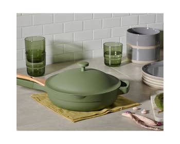 the green always pan on a kitchen counter surrounded by cups, bowls, and plates