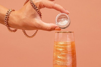 person pouring glitter into a champagne glass with an orange drink that looks sparkly