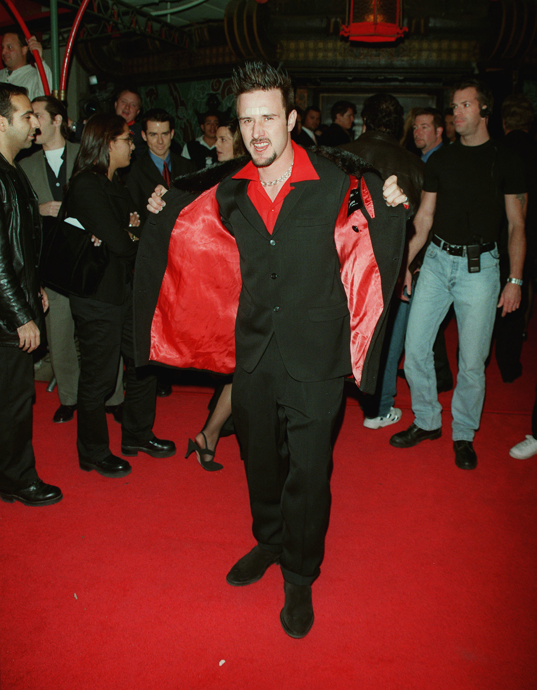 David Arquette in a black suit with a red collard shirt and a Band-Aid on his head