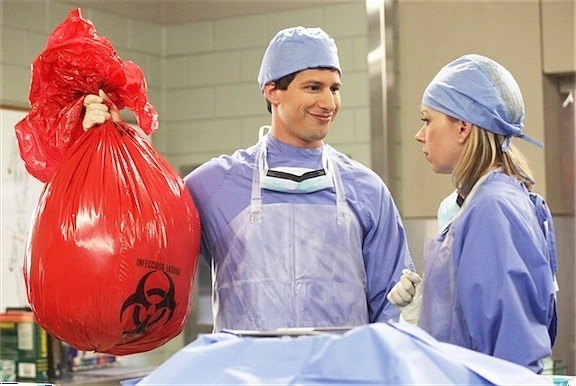 Jake is holding up a red bag and smiling towards another character. (Their both in hospital scrubs)