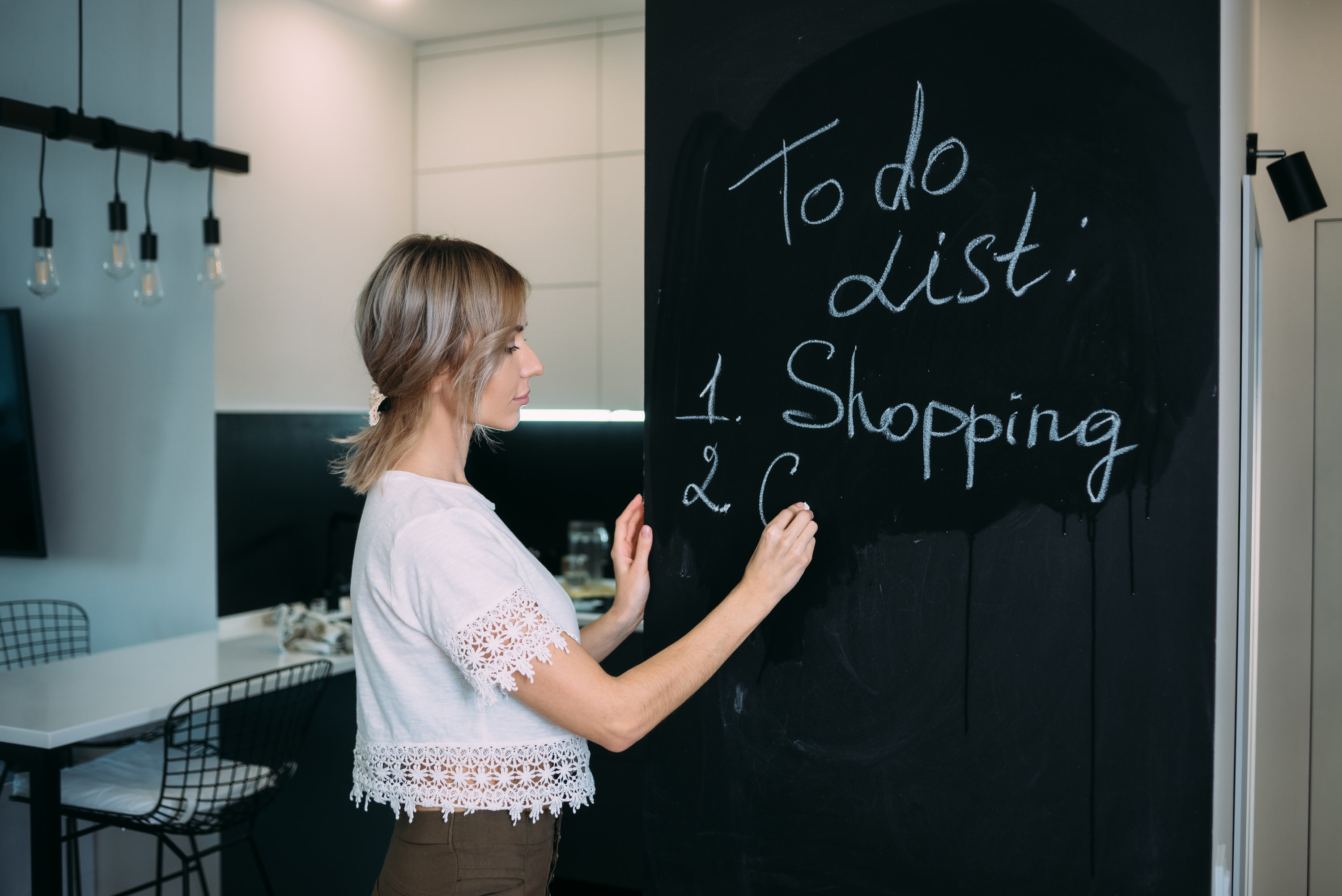 A woman stands in a kitchen, writing in chalk on a large blackboard wall