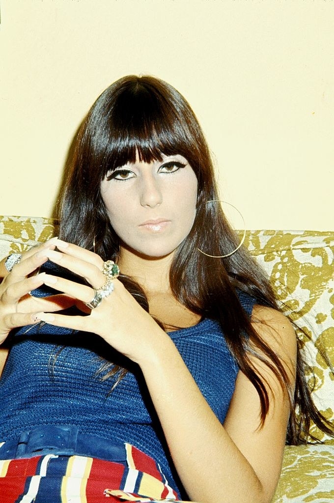 Cher in 1968 sitting on a couch with her hands crossed and wearing dramatic eye makeup