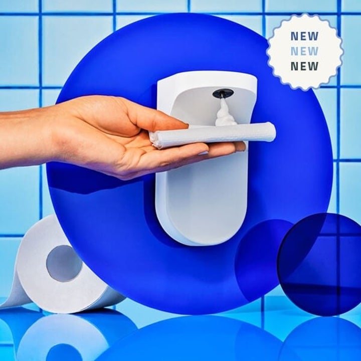 a white wall-mounted foam dispenser with a hand holding a piece of toilet paper underneath