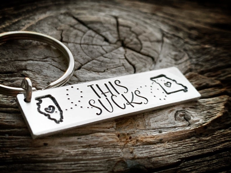 Silver keychain that says &quot;this sucks&quot; with state illustrations on it