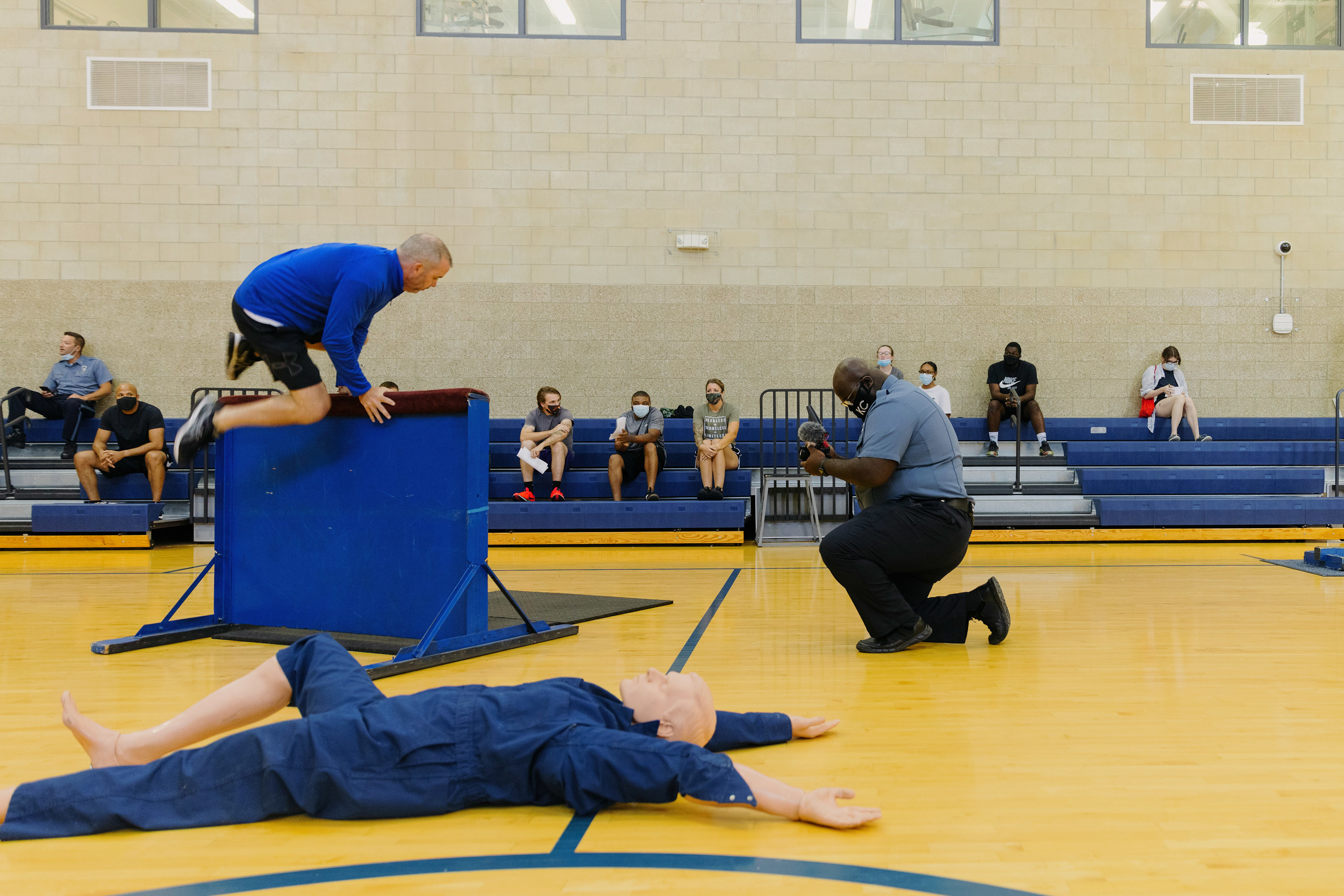 In a school gymnasium,  a man kneels and films another man hopping over a barricade