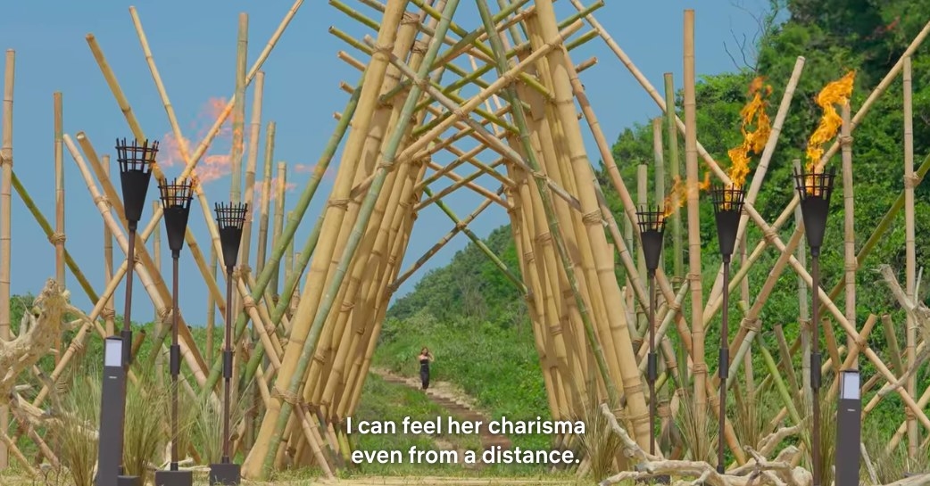 Torches are lit on both sides of a bamboo fixture, a woman approaches from a path in the distance