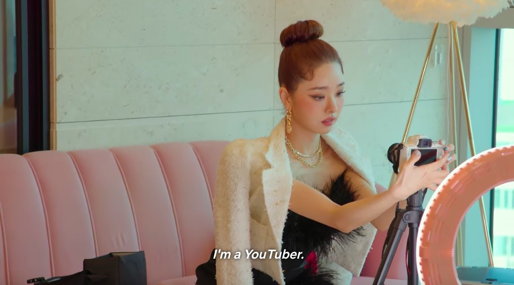 Ji-a sits on a pink velvet couch all dressed up, focusing a camera on herself