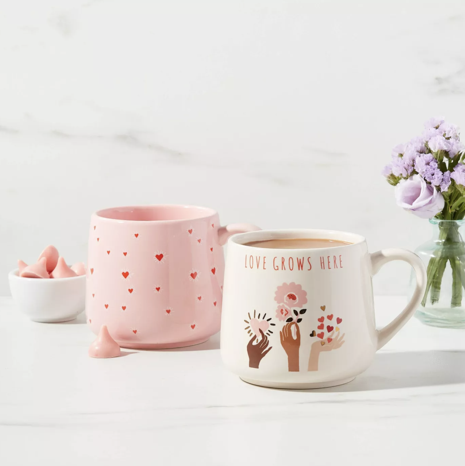 A pink mug with red hearts and a white mug with three hands holding flowers and the words &quot;love grows here&quot;