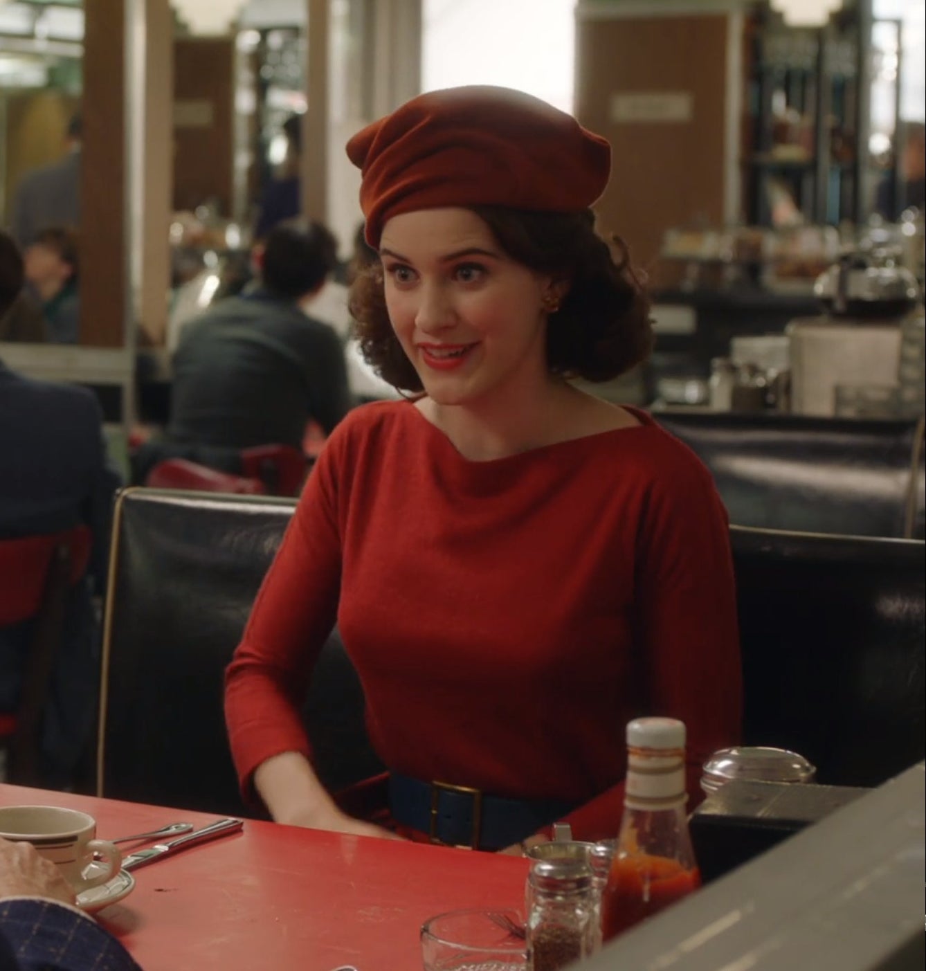 Screenshot from Mrs. Maisel shows Midge in a red dress sitting in a booth