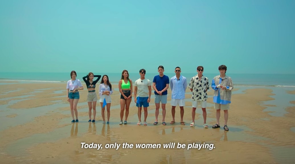 The singles stand in a line on the beach as the announcer says only women will be playing today