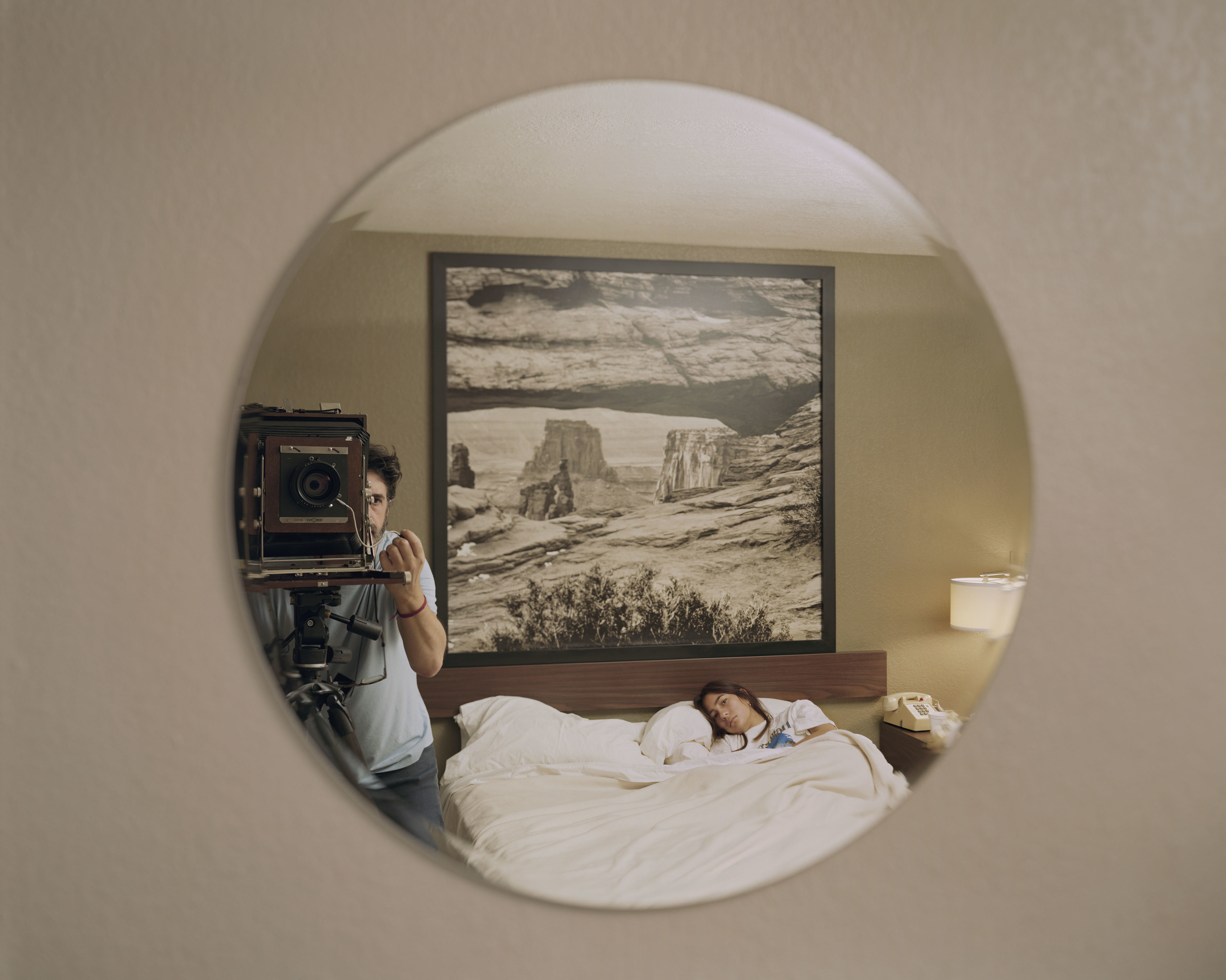 Alec Soth with a large format camera in a round mirror reflecting him and his daughter in bed