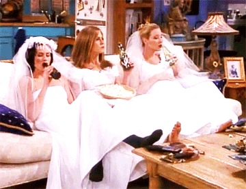 Monica, Rachel, and Phoebe from Friends, wearing wedding dresses and drinking beer on the couch