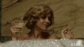 Christina&#x27;s character holding a martini in one hand and grabbing a bottle of clear liquor in the other as she sits in a bubble bath