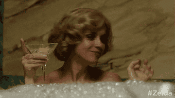 Christina's character holding a martini in one hand and grabbing a bottle of clear liquor in the other as she sits in a bubble bath