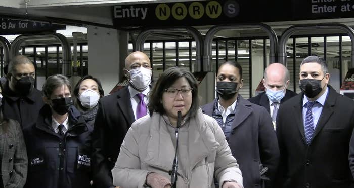 A woman stands at a microphone inside a subway station with various people, including NYC Mayor Eric Adams, standing behind her and wearing masks