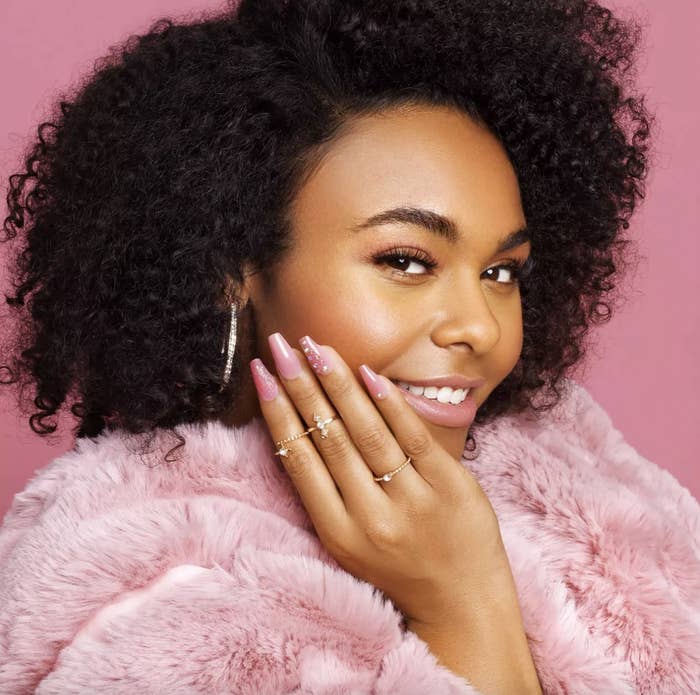 A person with curly hair, a pink fur coat and pink bedazzled nails