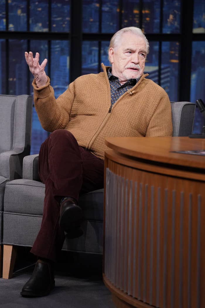 Cox gestures his hand out while sitting on a couch at a late-night show
